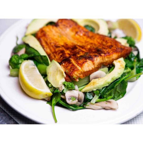 Grilled salmon fillet with avocado, broccoli, mushrooms, spinach, and lemon wedges on a white plate.