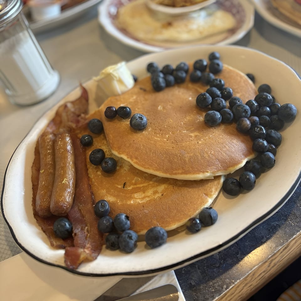 Diner breakfast plate with blueberry pancakes, bacon, sausage, and butter.